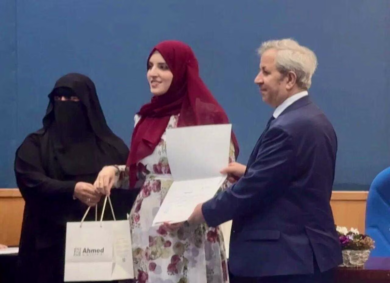 One Man and One Women from Head of Arabic Department at an award ceremony handing a certificate to Rawan who is wearing a floral dress and red hijab.