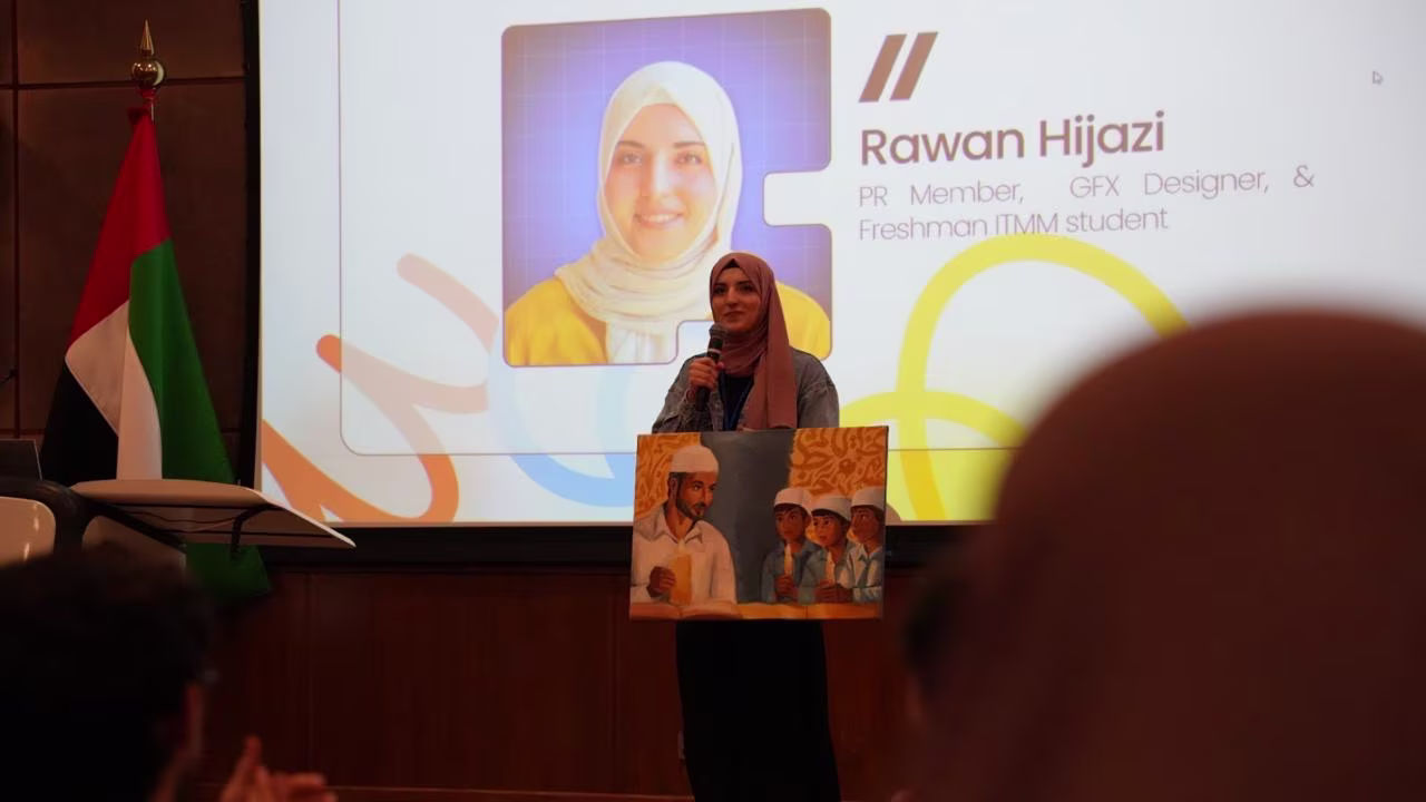Rawan wearing a pink hijab stands on a stage, holding a microphone and a painting depicting a man and three children, with a presentation slide behind her showing her photo and name, Rawan Hijazi, along with her title as PR Member, GFX Designer, & Freshman ITMM student. A UAE flag is visible to the left.