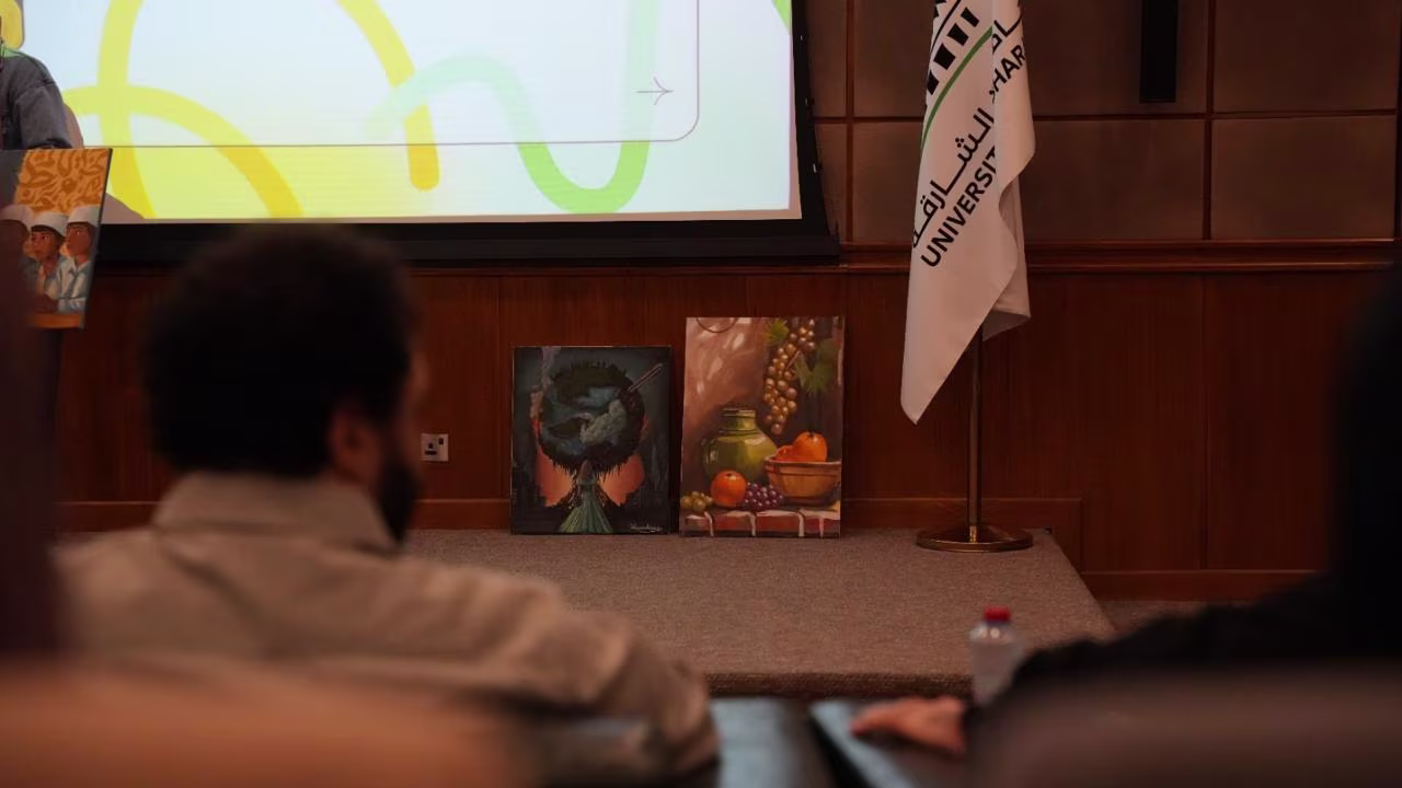 Audience members seated facing a stage with two paintings displayed on the floor and a flag on a stand in a wood-paneled room.