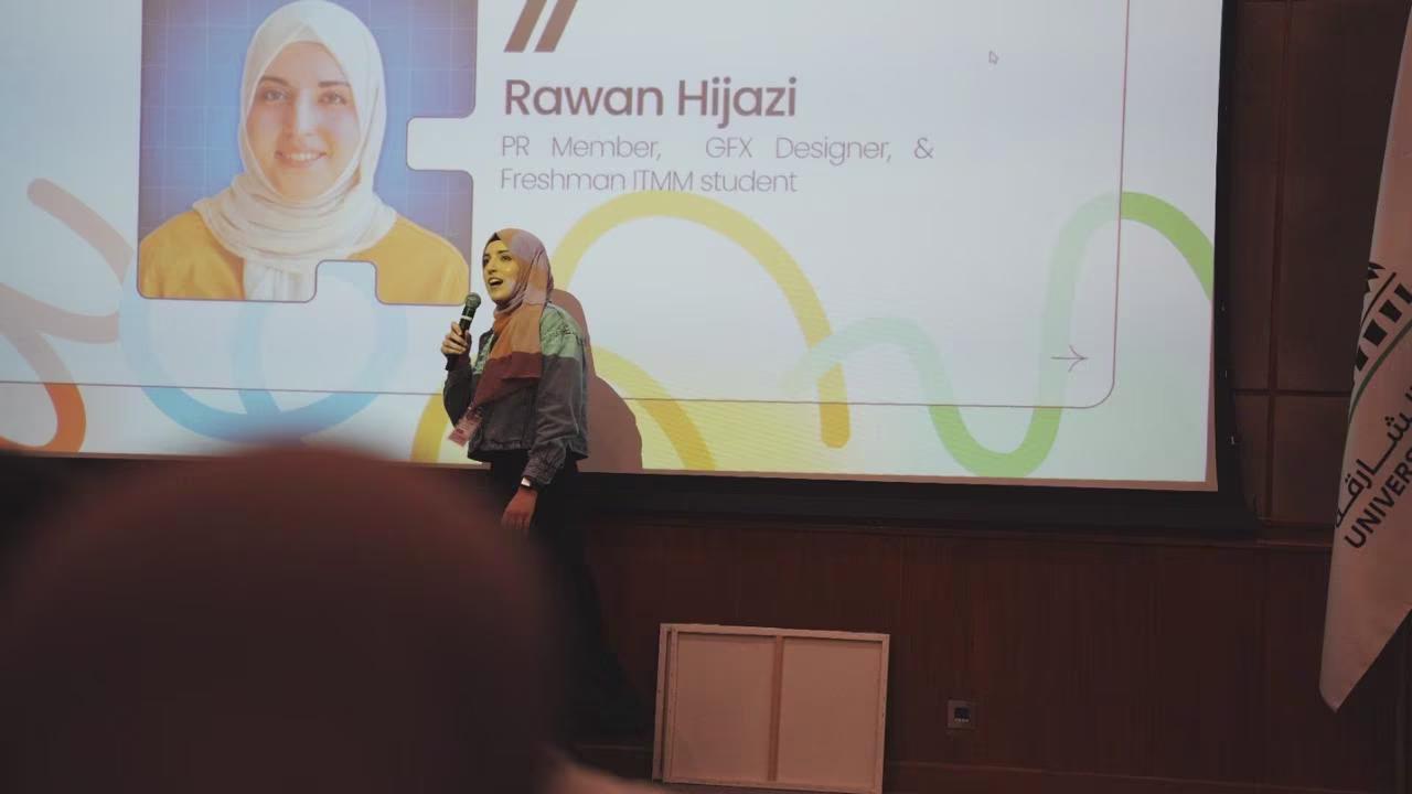 Rawan wearing a hijab and holding a microphone is speaking in front of a projected slide that shows her name, Rawan Hijazi, and her roles as PR member, GFX designer, and ITMM freshman student.