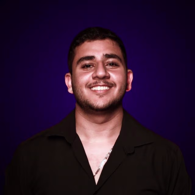 Portrait of a smiling man with short dark hair and trimmed beard wearing a black shirt against a dark blue background.